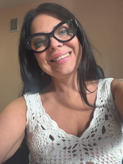 A woman is posing for a photograph. She has dark hair and is wearing glasses and a white, crocheted top. She is smiling at the camera. The background is a plain wall. There is no indication of Murmansk, Russia in the image.