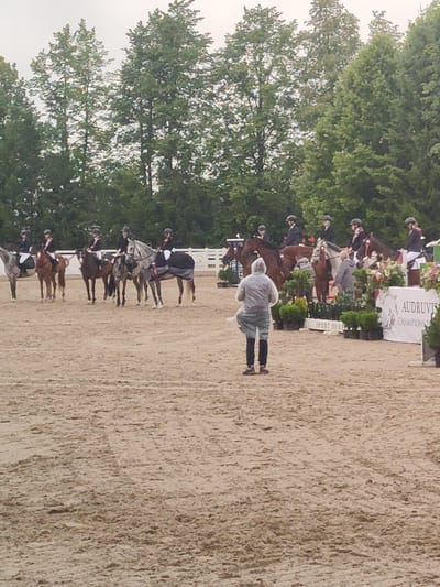 In the city of Ziniūnai, Lithuania, an outdoor equestrian event is underway. The foreground shows a sandy arena where a person in a light-colored, hooded rain poncho is standing. Behind them, a line of horses and riders are positioned. The riders are wearing helmets and riding attire. Several horses are adorned with blankets. In the background, a white fence and banners are visible. One banner reads "AUDROVIS CHAMPIONSHIP". Beyond the fence, a line of trees forms the backdrop against a cloudy sky. The environment suggests a competition or exhibition of equestrian skills.