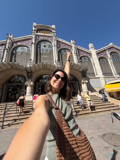 This is a selfie taken outdoors, with the subject's right arm and shoulder in the foreground, holding a brown woven bag. The subject, a woman wearing sunglasses and a green top, is smiling at the camera. Her left arm is raised, pointing towards the architectural facade of a large building behind her. The building appears to be a market, with ornate details, large arched windows, and a yellow sign that reads "HORCHATA CHURROS".
Several other people are visible on a wide set of stairs leading up to the building's entrance. They are casually dressed, suggesting a daytime activity. The sky is a clear, bright blue, indicating fair weather and likely a sunny day. The architecture and the presence of "HORCHATA" on the sign strongly suggest the location is in Valencia or its surrounding areas, specifically Alboraia/Alboraya, known for its horchata. The overall mood is cheerful and tourist-oriented.