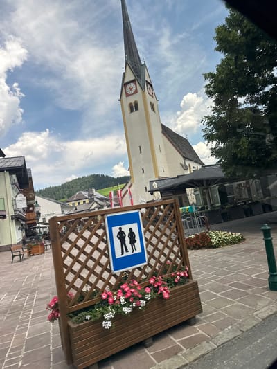 The image captures a street scene in the city of Au, Austria, on a cloudy day. In the foreground, a wooden planter box filled with pink and white flowers stands on a paved square. A blue sign with silhouettes of a man and a woman, indicating a restroom, is mounted on the planter. Behind the planter, a church with a tall, pointed steeple dominates the skyline. The church is white with yellow trim and features two clocks on its tower. To the left of the church, a row of buildings with traditional European architecture lines the street, including a building with a sign that reads "Gasthof Hirschbichler". Trees and hills covered in lush green vegetation are visible in the background. Outdoor seating areas with umbrellas suggest the presence of cafes or restaurants. The overall scene depicts a charming town square with a mix of historical architecture and natural beauty.