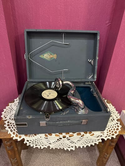 A vintage portable record player is open and displayed on a crocheted doily atop a wooden side table. The record player is a dark gray color with a silver tone horn and needle. A black vinyl record is on the turntable. The background is a textured, deep pink wall. The scene is indoors. There are no people present. The location is specified as Fort-Şevtşenko, Kazakhstan.