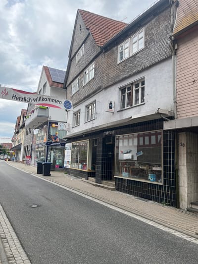 The image depicts a street scene in Neustadt, Germany, under a cloudy sky. A banner that reads "Herzlich willkommen" (Welcome) is strung across the street. The scene features buildings with traditional German architecture, including steeply pitched roofs covered in wooden shingles and tile.

On the right side of the street, a building with a light gray facade has a shopfront with a black tiled exterior and large display windows. The windows display various items, including what appear to be model airplanes and boxes. The building's entrance is marked with the number "20".

To the left of this building, another shopfront is visible, featuring a blue and white facade with the text "Photovoltaik Elektromobilität" and an image of a worker in a hard hat. This shop appears to be selling solar power and electric mobility solutions. Two dark-colored trash bins are placed in front of this shop.

Further down the street on the left, more buildings and shopfronts are visible, suggesting a commercial area. A red tiled roof and solar panels are visible on one of the buildings. The street itself is paved with asphalt and bordered by a sidewalk made of cobblestones. The overall impression is of a quiet, European town street.
