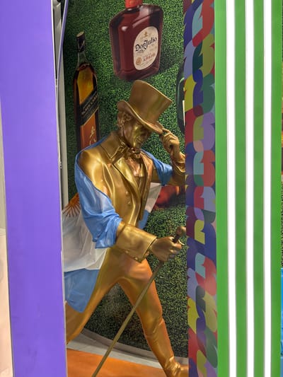 In Ezeiza, Argentina, a golden statue of a man in a top hat and waistcoat stands to the right of the frame, holding a cane and tipping his hat. He is draped in a light blue fabric with a sun emblem, resembling the Argentine flag. Behind him, a wall displays images of two liquor bottles: Johnnie Walker Black Label and Don Julio Añejo, set against a green, textured background. To the right of the statue, a vertical column features a colorful, abstract pattern of interlocking shapes in shades of red, blue, yellow, green, and purple. Further to the right, a bright green panel with three vertical white illuminated bars completes the scene. A portion of a purple panel is visible on the far left of the image.