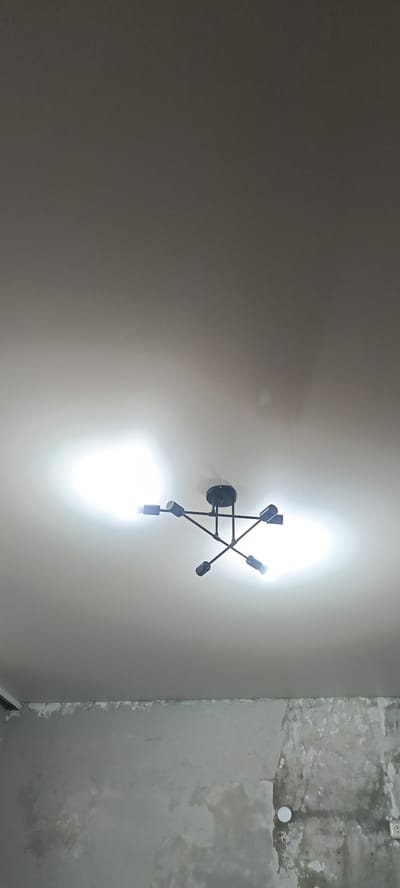 The image shows a modern, black spider-like chandelier hanging from a white ceiling. The chandelier has multiple arms extending outwards, each ending in a light fixture. The lights are on and casting a bright glow on the ceiling. Below the ceiling, a wall is visible, painted in a muted grey color. The wall appears to be unfinished or in disrepair, with patches of peeling paint and textured surfaces. There is a white circular fixture mounted on the wall, possibly an electrical outlet or a light switch. The overall scene suggests an indoor setting, likely a room with unfinished renovations. The image does not contain any discernible information related to the city of Хохлы, Russia.