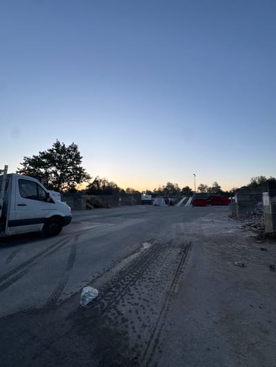 This image captures a static scene at what appears to be an industrial yard or construction material handling facility in Émerainville, France, during either dusk or dawn. The sky, a clear gradient from deep blue overhead to a soft orange and yellow near the horizon, suggests the transition of day to night or vice-versa.
In the foreground, a dark, concrete or asphalt ground shows numerous tire tracks and wet patches, possibly from recent rain or spills. A white plastic bag is visible on the ground to the right. A white utility truck, likely a van, is parked on the right side of the frame, facing away. On its side, the text "Construction Renovation" is clearly legible.
Further into the midground, a large, leafy tree stands prominently. Behind it and stretching along the horizon, several red industrial containers are stacked, along with piles of concrete blocks and other construction debris, indicating the nature of the site. A street lamp stands tall between the containers and the tree. In the background, more trees and a building or wall are visible. No people are present, and no specific activity is taking place, giving the scene a quiet, still atmosphere.