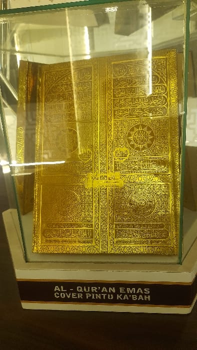 An indoor scene in Glantengan, Indonesia, displays a rectangular, golden-colored object vertically within a clear glass display case. The object, which appears to be a decorative replica or cover, is extensively adorned with intricate Arabic calligraphy and geometric patterns, resembling an ornate door. It rests on a multi-tiered base, primarily white with a dark brown band at its lower edge. The brown band features white capital lettering that reads "AL - QUR'AN EMAS COVER PINTU KA'BAH". Reflections from an unseen light source are visible on the glass enclosure. No individuals are present in the frame.