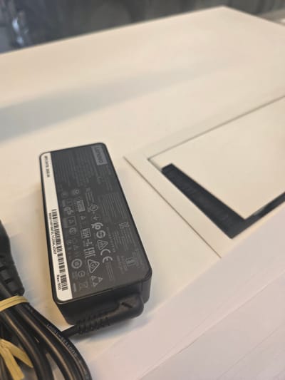 The image shows a Lenovo laptop power adapter with its cable on a white surface. The adapter is black and has various safety and compliance markings, including "Lenovo," "Max 65W," and "MPO DATE: 2020.04." The adapter has multiple regulatory certifications like cULus, NOM, SACE, and EAC. The cable is black and partially coiled. The surface the items are on appears to be a white laminate. No people, subjects, or actions are visible. The location in Sint-Pieters-Leeuw, Belgium is not discernible from the given image.