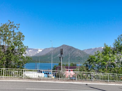 The image depicts a panoramic view of a roadside area in Kirovsk, Russia, with a backdrop of majestic mountains and a clear blue sky. In the foreground, a paved road with white lane markings is visible, suggesting a transportation route. A white metal fence runs along the edge of the road, separating it from a strip of vegetation and some buildings. Beyond the fence, a body of water, likely a lake or reservoir, stretches across the mid-ground.

The landscape is characterized by a prominent mountain range in the distance, with snow-capped peaks that indicate a high altitude or a cooler climate. The slopes of the mountains are covered with green trees and vegetation, transitioning to a rocky, barren appearance at higher elevations. Several utility poles and a pylon are scattered throughout the scene, hinting at infrastructure development in the area.

A few buildings with red and pink roofs are nestled among the trees near the water's edge, suggesting a small settlement or residential area. The overall impression is one of a picturesque natural setting with human presence, typical of a town or city located in a mountainous region. The bright sunlight and clear sky suggest a pleasant day for outdoor activities.