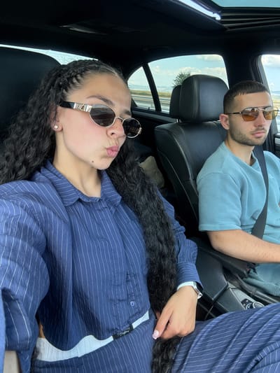 Two individuals, a woman and a man, are seated in the front seats of a vehicle, likely a car, traveling on a highway. The woman, in the foreground on the left, is leaning towards the camera, making a kissing face. She has long, dark, curly hair styled in braids and is wearing a blue pinstripe shirt and matching pants, along with oval-shaped sunglasses and a diamond stud earring. The man, on the right, is looking forward, wearing a light blue ribbed t-shirt and sunglasses with orange lenses. He has short, dark hair and stubble. The interior of the car appears to be dark leather. The background through the windows shows a landscape with rolling hills, trees, and a cloudy sky, suggesting an outdoor, possibly rural setting. The overall impression is a candid moment captured during a road trip. The location is identified as Polsko Padarevo, Bulgaria.