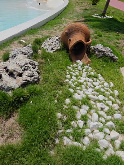 An overhead shot captures an outdoor scene, possibly a park or garden, with a pool on the left. A broken terracotta amphora lies on its side in the grass, with its contents, white stones, scattered around it, forming a stream-like pattern. Several large, rough rocks are strategically placed throughout the grassy area. A hint of a palm tree and a red running track can be seen in the background, suggesting a recreational area in Cancún, Mexico.
