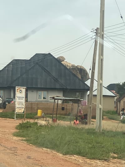 The image shows a scene in the city of DU, Nigeria. In the foreground, a dirt road leads towards a grassy area with a white goat standing amongst the vegetation. Behind the goat, a wooden table holds a yellow plastic container. To the left of the table, a sign for "THE REDEEMED CHRISTIAN CHURCH OF GOD" is visible.
Further back, a house with a dark, sloped roof is prominent. Adjacent to this house, several other buildings with corrugated metal roofs can be seen. Several people are gathered under a simple wooden shelter, with clothing hanging on a line. A few individuals, including children, are visible in the mid-ground and background of the scene. Electrical poles and wires stretch across the sky. A large rock formation is situated behind the main house. The sky is overcast.