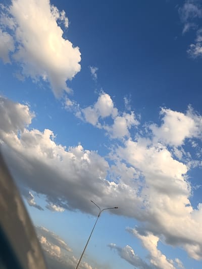 A low-angle, full shot captures a bright blue sky dotted with fluffy white and gray clouds. A single streetlamp with a curved arm and a small fixture is visible in the lower right quadrant, extending upwards and to the left. The base of what appears to be a vehicle or structure is visible along the left edge of the frame. The background appears to be a landscape with hints of mountains or hills below the clouds. The scene is set in Unknown, Kazakhstan.