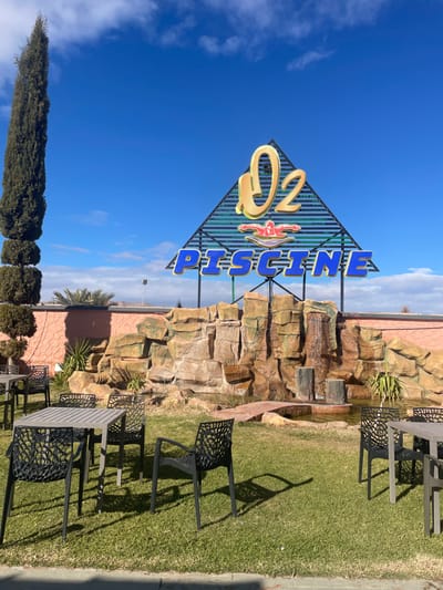 The image displays an outdoor leisure area identified as "O2 Piscine" in Lkheng, Morocco, under a clear blue sky with sparse clouds. In the foreground, a vibrant green lawn is set with multiple dark grey outdoor tables featuring slatted tops and accompanying chairs with intricate, open-weave backrests. Strong sunlight from the right casts crisp shadows of these chairs onto the grass.

The midground is dominated by a man-made rock formation designed as a waterfall, with water visibly cascading into a small pool at its base. The rockwork exhibits varied earthy tones of brown, ochre, and green. A reddish-brown, stone-like path or bridge traverses a section of the water. Behind this feature, a peach-colored rendered wall spans the scene. To the far left, a tall, slender cypress tree is prominently featured, trimmed with two distinct, lower spherical sections.

In the background, a large, triangular A-frame sign stands above the rock formation, displaying "O2" in prominent gold lettering at the apex, a stylized red swimmer figure below it, and "PISCINE" in bold blue lettering at the base. The sign structure appears to be made of dark metal latticework. Beyond the wall and sign, an arid landscape with distant low hills and sparse vegetation, including a lone palm tree, is visible under the expansive blue sky. A street light pole is discernible behind the sign. No individuals are present in the scene.