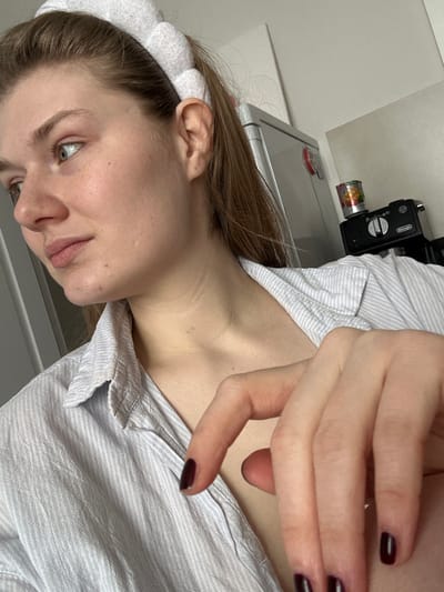 An indoor, domestic scene is depicted in Nikol'skii, Russia, featuring a young adult female from the chest up. She has fair skin, light brown hair pulled back with a segmented white terry cloth headband, and is wearing a light-striped, collared shirt unbuttoned at the neckline. Her face is presented in profile, gazing towards the left of the frame, revealing light-colored eyes and natural facial features, including minor blemishes on her left cheek and jawline. Her left hand is visible in the foreground, with nails painted a dark burgundy color. The background consists of a light-colored wall. A silver-toned refrigerator or cabinet is partially visible on the left. To the right, a black Delonghi coffee machine rests on a counter, topped with a colorful tin can. A small red object is affixed to the wall near the refrigerator.