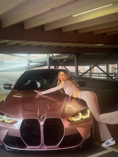 A woman with blonde hair and fair skin is posing on the hood of a pink BMW M4 coupe. She is wearing a light-colored, fitted long-sleeved shirt and matching pants. Her right arm is extended, resting on the car's hood, and her body is arched. The car is parked in what appears to be a multi-story parking garage, with concrete floors and overhead structures visible. The lighting in the garage is artificial, casting a warm glow on the scene. The location is identified as Kołobrzeg, Poland.