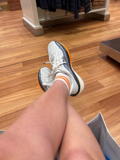 This is a close-up, high-angle, eye-level shot of a person's legs and feet, positioned on a polished wooden floor. The person is wearing white athletic shoes with orange and black accents, and white socks with orange stripes. The shoes appear to be brand new, with laces neatly tied and the soles showing no signs of wear. The person's legs are crossed, with the right leg resting over the left. A white shopping bag is visible on the right side of the image, its edges blurred. In the background, on the left, a display of folded clothing items can be seen, suggesting the image was taken in a retail environment. The lighting is bright and even, indicating it is likely daytime, and the overall mood is neutral and observational. No text is visible in the image.
