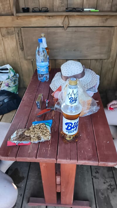 A brown wooden picnic table is set up outdoors, with two people partially visible at the bottom of the frame. On the table, a bottle of "Big Urban Beer," a bottle of water, a pack of cigarettes, a lighter, a knife, a small figurine, a straw hat, a bag of snacks, and some sliced meat are laid out. The scene is illuminated by natural daylight. The location is the village of Kiriushkino, Russia.