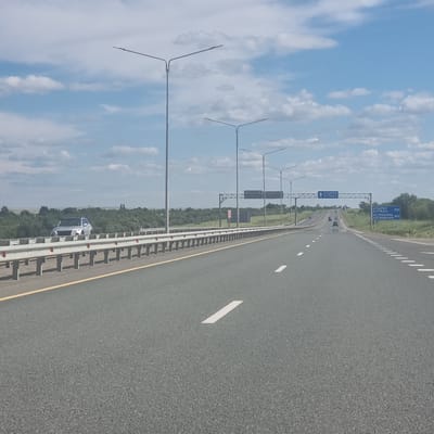 The image shows a highway leading towards a distant horizon under a partly cloudy sky. Tall lampposts line the right side of the road. A silver SUV is visible in the distance on the left side of the road, behind a guardrail. A blue road sign is visible on the right side of the road indicating directions and distances to Atyrau (552 km) and Astrakhan (902 km). The road is asphalt with clear lane markings. The environment appears rural with vegetation and trees visible in the background. The location is identified as the highway near Alğa, Kazakhstan.