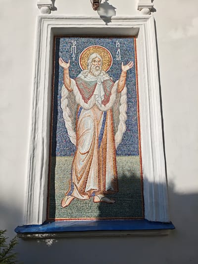 The image shows a mosaic depicting a standing figure of a bearded man with a halo, possibly a religious figure. The mosaic is set in a white framed niche on a light-colored building exterior. The background of the mosaic is a dense pattern of blue and white tiles. The mosaic itself features earth-toned robes and white hair. Above the figure are Cyrillic characters. The building's exterior is plain and white with simple architectural details. The base of the niche is painted blue. The location is Мамонтово, Russia.