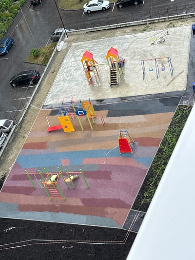 The image shows an overhead view of a newly constructed playground in the city of Засечное, Russia. The playground is situated on a multi-colored, impact-absorbing surface with sections of gray concrete. To the left of the playground, a paved road with painted parking spaces is visible, with several cars parked, including a blue hatchback and a black sedan. Rain is falling, and the road and playground surfaces are wet.
The playground equipment includes a climbing structure with a red slide, a swing set with two blue swings, and a separate, smaller climbing structure with a red base and netting. Further to the right, another section of the playground features a climbing frame with green poles and a red ladder, and two yellow seesaws. The perimeter of the playground area is marked with white and red striped caution tape. Adjacent to the playground is a green planting strip with young plants, and beyond that, a white building structure.