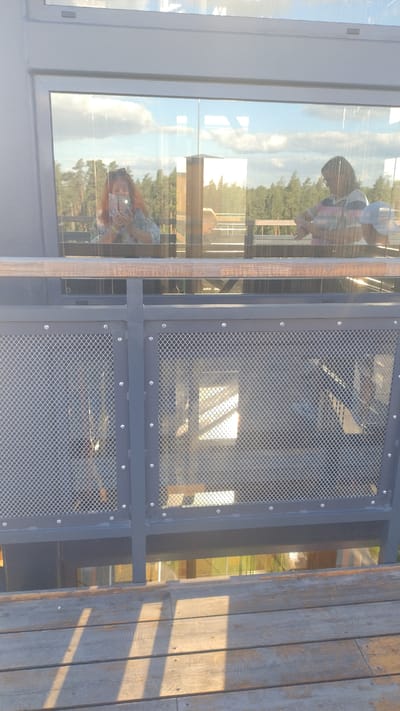 In Tērvete, Latvia, a woman with red hair is taking a selfie in front of a large glass window. She is holding a smartphone and wearing a patterned shirt. Next to her, a person with dark hair and glasses, wearing a striped shirt, is visible. The reflection in the window shows a forest and a sky with clouds. Below the window, a wooden railing and a metal fence with a mesh pattern are visible. The scene appears to be outdoors, possibly on an elevated platform or walkway.