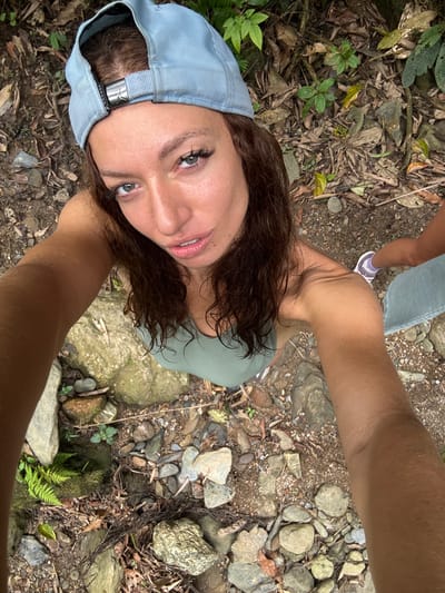 The image shows a first-person perspective selfie taken by a woman in a natural environment. The foreground features the woman's face wearing a light blue baseball cap, and she appears to be looking directly into the camera. Her arms extend towards the camera, framing the shot. In the midground, the background shows a rocky, foliage-covered area, indicative of a natural setting. A portion of another person's leg wearing jeans and a sneaker is partially visible in the top left corner. The ground is covered with rocks, leaves, and small plants. Based on the location context, it is possible that the photo was taken in Suối Giàng, Vietnam.
