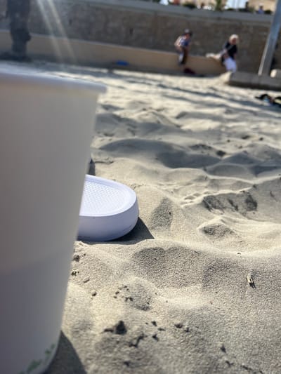 The image shows a close-up view of a white shoe partially buried in sand, possibly at a beach. The sand is light in color and has a grainy texture. In the background, out of focus, there are some people walking along a walkway or path. Given the location of Peguera, Spain, it can be assumed the location is likely a beach, and the people are likely tourists enjoying the coastal scenery. The shoe and the container partially obstruct the lower part of the image, giving a first-person perspective. The container appears to be an item left on the ground.