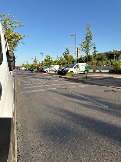 An eye-level shot captures a sunlit urban scene in Getafe, Spain, dominated by a clear, bright blue sky. In the foreground, the right side of a white van is partially visible, its side mirror also in view. The van appears to be parked along a street or parking lot.
The asphalt road stretches into the distance, marked with white lines and painted speed limit signs that read "20" within squares. Several trees are planted along the side of the road, their green leaves contrasting with the asphalt and blue sky. Beyond the trees, more parked vehicles, including vans and cars, are visible. One white van has the green "Orona" logo and text that reads "going closer." In the background, construction cranes suggest ongoing development. Buildings can be seen further in the distance, and other street lights are present. The overall environment suggests a commercial or industrial area during daytime.