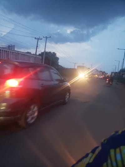 The image depicts a road scene in Ida, Nigeria, at dusk or dawn. The sky is a gradient of blue, with dark clouds on the left side of the frame. Power lines and utility poles are visible against the sky. A road stretches into the distance, with vehicles present on it. Bright headlights suggest traffic moving towards the camera's position. A red car is partially in the frame, occupying the lower central part of the image. The edge of a building or structure is present on the left side, alongside the road. A partially visible subject is also present on the upper right, with colorful patterns on fabric.