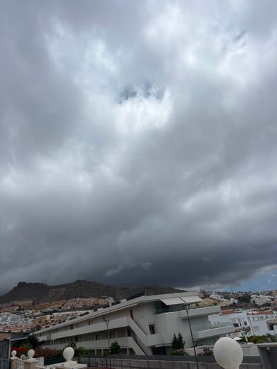 The image shows a cloudy sky over a city on a hillside, possibly Miraverde, Spain. The clouds are dark and heavy, suggesting an impending storm. The buildings in the city are clustered together on the steep slope, with a mix of white and colored facades. In the foreground, there is a white building with balconies and an awning. A white spherical object is visible on the right side of the frame.