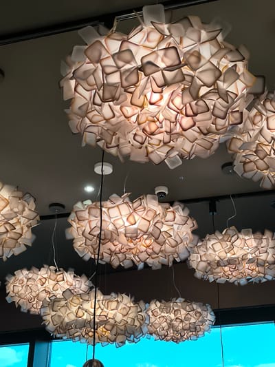 The image shows a low-angle view of a ceiling with multiple light fixtures. The fixtures are ornate, spherical, and made up of numerous small, square-shaped pieces that appear to be translucent or semi-translucent. The pieces are a mix of light and dark shades, possibly due to shadows and the light source within the fixtures. A security camera is also visible on the ceiling. A window can be seen in the bottom left corner, showing a view of a bright blue sky. The location is Schwechat, Austria, as it can be inferred from the surroundings.