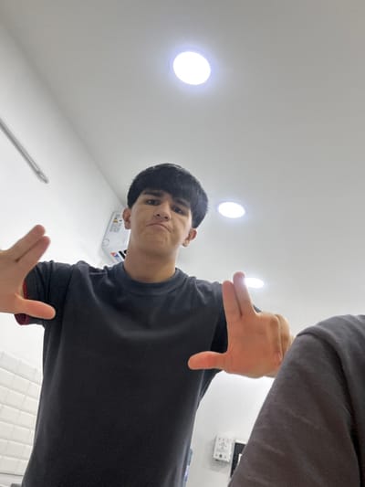 A low-angle shot features a person making a finger-frame gesture. The person is centered, looking down at the camera with a neutral expression. Their dark hair is neatly styled, and they are wearing a dark t-shirt. The background is a white ceiling with recessed lighting. The angle suggests the photo was taken from below, looking up at the subject. Parts of another person's arm are visible in the upper right.