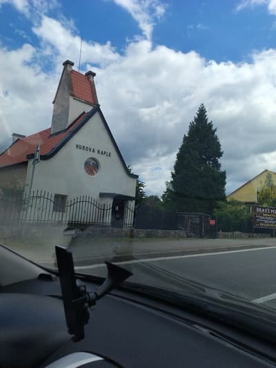 The image captures a view of a building identified as "HUSOVA KAPLE" in Bernartice, Czechia. The building has a light-colored facade with a red tiled roof and a prominent, angular chimney. A circular window with a spoked design is visible on the front of the building. In front of the chapel, there is a black wrought-iron fence and some foliage. To the right of the chapel, a tall evergreen tree stands prominently. Further to the right, a yellow building is partially visible, and a large banner with text, including "HRAVE PER...", is displayed on the fence. The scene is viewed from the interior of a vehicle, with a car mount for a phone visible on the dashboard in the foreground. The sky above is blue with scattered white clouds. A road is present in the foreground, suggesting the photograph was taken from a moving vehicle.
