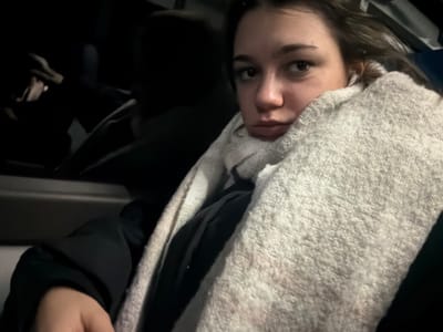 A dimly lit, close-up image captures a young woman seated inside what appears to be a vehicle, likely a car, in Obermodern-Zutzendorf, France. She is the primary subject, looking towards the camera with a somewhat somber, tired, or slightly pouting expression. Her dark brown hair is visible around her face, and she wears a white earbud in her right ear.

She is bundled warmly in a thick, light-colored, possibly off-white or light grey, knitted scarf or blanket that covers her neck and shoulders. Underneath, a dark-colored jacket or coat is visible. The immediate surroundings suggest the interior of a car, with elements like a seat and a door panel or armrest subtly visible in the low light.

The scene is predominantly dark, indicating that it is likely nighttime or that the vehicle's interior is very dimly lit. In the deep shadows to the left, the hands of another individual can be seen, apparently holding and interacting with a smartphone or similar device. The activity suggests either traveling or waiting. The overall mood conveyed by the woman's expression and the dark setting is one of quiet contemplation or weary repose. No text is visible in the image.
