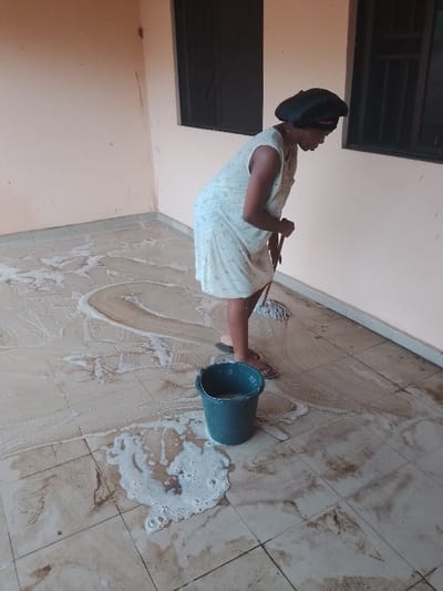 An adult female is depicted actively mopping a tiled floor in Ufuma, Nigeria, within an interior or covered exterior space featuring light peach-colored walls and dark-framed windows. She wears a light-colored, patterned sleeveless dress, dark flip-flops, and a black head bonnet with a red band. The individual is bent at the waist, holding a mop with a dark handle and light-colored head, directing it across the floor. A dark green bucket, approximately half-filled with foamy liquid, sits on the floor to her left. The beige-tiled floor is extensively covered with white, frothy soap solution, visible dirt, and streaks indicative of ongoing mopping. Spilled suds are also present around the bucket.