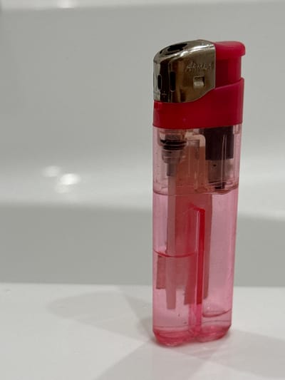 A pink disposable lighter with a silver metal top sits upright on a white surface, possibly a sink or countertop. The lighter contains clear liquid fuel, with a small amount of pink coloration. The surface appears clean, with subtle reflections of light. The background is a soft, out-of-focus gray, suggesting an indoor setting. There are no people visible in the image. This image was taken in Kamensk-Uralsky, Russia.