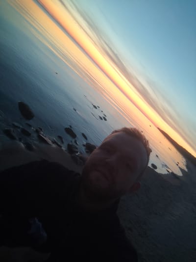The image is a tilted, dark, and grainy selfie taken outdoors at sunset. A man's face is visible in the lower right corner, with his head angled upwards. He has fair skin, light hair, and is wearing a dark t-shirt. The background features a serene body of water reflecting the orange and yellow hues of the setting sun, with a clear blue sky above. The shore is rocky and appears to be made of sand and pebbles. The overall impression is a candid moment captured during a tranquil evening by the water.