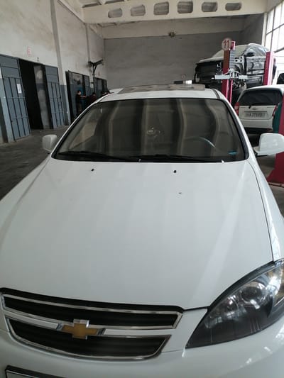 The image shows the interior of a car repair shop in Juma, Uzbekistan. A white Chevrolet car is parked in the foreground, facing the camera. In the background, several garage doors are visible, some open and some closed. A car is suspended on a lift in the middle background. Three men are standing near one of the open garage doors, appearing to be in conversation or working on something. The overall environment appears to be a functional workshop.