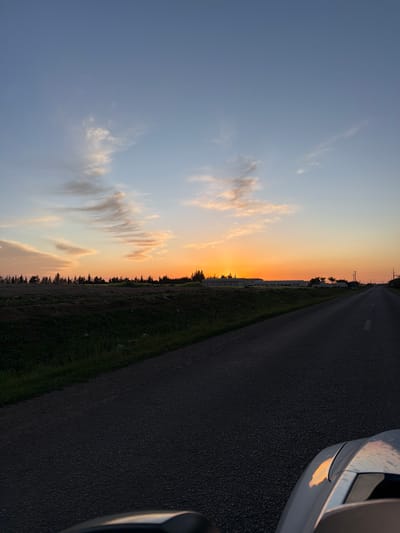 The scene depicts a rural landscape at sunset near Sabaa Aiyoun, Morocco. A two-lane asphalt road occupies the center and right foreground, extending into the distance with a faded white broken centerline. To the left of the road, a strip of dark green grass borders disturbed earth. Further left, a dense line of dark coniferous trees forms a silhouette against the horizon. Behind the trees in the midground, a cluster of light-colored buildings with dark roofs is visible. The sky transitions from a vibrant orange at the horizon, where the sun has nearly set, through lighter oranges and yellows, into a pale blue and then a deeper blue overhead. Wispy, elongated clouds are present, illuminated by the setting sun. Power poles with overhead lines run parallel to the road on the right side of the frame. The lower right corner of the image shows the front-right fender and side mirror of a light-colored vehicle, reflecting the orange hues of the sunset, indicating the perspective is from a vehicle on the road. No individuals are visible.