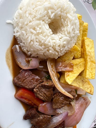 A close-up overhead shot shows a plate of Lomo Saltado, a Peruvian dish, at a restaurant in San Vicente de Azpitia, Peru. The dish consists of a mound of white rice, a pile of golden french fries, and stir-fried beef with onions and tomatoes in a savory sauce. The lighting is natural and appears to be from a window, casting soft shadows on the food. The plate is white and has a slight rim. The background is out of focus and appears to be a table with a patterned surface and a faint glimpse of a wine glass.