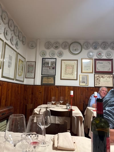 The image depicts the interior of a restaurant in San Biagio di Callalta, Italy, with a focus on the wall decorations and a dining table in the foreground. The walls are adorned with numerous decorative plates, many featuring botanical illustrations. Several framed documents and newspapers are also visible on the wall, adding to the establishment's ambiance.
In the foreground, a dining table is set with white tablecloths, silverware, wine glasses, and a bottle of red wine. Two men are seated at tables in the background. One man, with graying hair, is wearing a blue shirt and is looking towards the camera. The other man is partially visible and wearing a denim shirt. The room has wooden paneled walls and a warm lighting that suggests an evening setting. The overall impression is of a cozy and traditional Italian dining establishment.