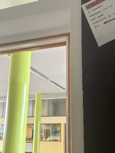 The image is taken from inside a building, looking out of a window, possibly in Kranj, Slovenia. A piece of white paper with bold lettering "VIKE..." and other text in Slovenian is partially visible on the top left corner. The window frame is white and has a dark wooden border around the glass. Outside the window, there are green horizontal pipes or beams in front of a yellow building with visible windows. Through the windows of the yellow building, what appear to be photographs or displays can be seen. Part of a white staircase structure with metal railings is visible through the window as well.