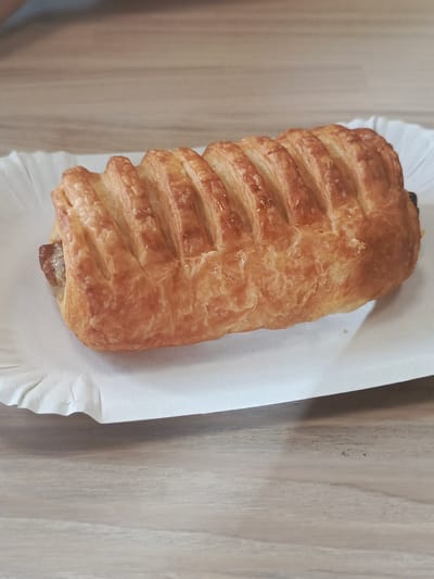 A close-up, eye-level shot shows a single, golden-brown pastry with a braided top layer, resting on a white paper tray. The pastry appears to be a sausage roll, with a dark filling partially visible at the left end. The pastry is positioned on a light-colored, wood-grained surface, possibly a table. The background is blurred and appears to be a continuation of the same wooden surface. There are no people or other objects clearly discernible in the image. The lighting is consistent and suggests an indoor environment. There is no information available to determine the location in Petrozavodsk, Russia, or any specific context for this image.