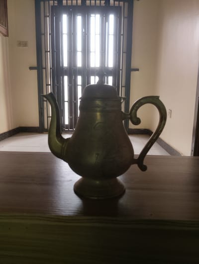 An antique brass teapot sits on a wooden surface in front of a closed metal gate with vertical bars. The gate is set against a wall, and sunlight streams through the gaps in the bars, creating vertical illuminated lines. The teapot is ornate, with a curved handle and a spout. The background wall is a light color, and there is a power outlet visible on the wall to the left of the gate. The floor appears to be tiled. The scene suggests an indoor setting, possibly a room or hallway with a window or door covered by a security gate. The image is taken from a medium-low angle, focusing on the teapot. The lighting is dim overall, with the brightest area being the light coming through the gate. The location is stated to be Alagbado, Nigeria.