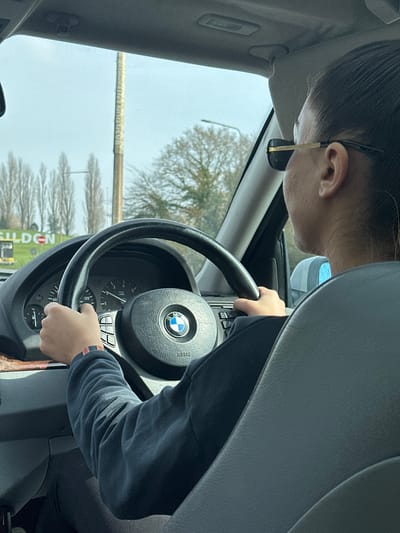 A first-person perspective photograph captures the interior of a vehicle and the external environment near Basildon, United Kingdom. A person, visible from the back of their head and right profile, occupies the driver's seat. They are wearing dark rectangular sunglasses with gold-colored arms and a dark long-sleeved garment. Their hands are positioned on a black BMW steering wheel, which features a central BMW logo and "AIRBAG" inscription, along with multifunction controls on both sides. A multi-colored striped wristband is present on the driver's left wrist. The vehicle's instrument cluster, partially visible, indicates a speed of approximately 20-30 mph. Through the windshield, a large green roadside sign displaying "BASILDON" in white capital letters is visible on the left, confirming the location. Adjacent to this sign, a tall, light-colored cylindrical pillar or monument with vertical text, including "BASILDON," stands prominently. The background features several bare deciduous trees, a streetlamp, and an overcast sky.