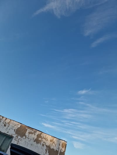 A low-angle shot captures a bright blue sky with scattered white cirrus clouds, stretching across the frame. In the upper right corner, the weathered facade of a building is visible. The building's surface is textured and peeling, revealing patches of rough material underneath a light-colored coat. A dark, curved edge of what appears to be a window frame or awning intrudes into the scene from the bottom right. A sliver of a green-framed window is also visible on the building's surface. The composition is dominated by the sky, suggesting an outdoor scene on a clear day. The building's aged appearance hints at a historical or older urban setting. There is no visible text in the image.