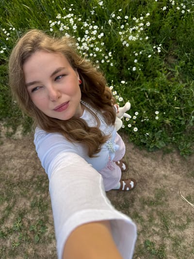 A young woman with long, wavy brown hair is lying on her back in a grassy field dotted with small white daisies. She is looking up at the camera, which is held by someone else. She has fair skin, blue eyes, and is wearing a white shirt with puffy sleeves and a pink and white striped skirt. Around her neck is a pearl necklace. She is also holding a small white stuffed animal. The background consists of more grass and some dry, bare earth, suggesting an outdoor setting, likely in a field or meadow. The lighting appears to be natural daylight, possibly late afternoon or early evening given the warm tones. The overall mood is peaceful and serene, with the woman's expression conveying a sense of calm and perhaps playfulness. The location is identified as Snovitsy, Russia.