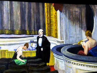 This painting, located in Reus, Spain, depicts an indoor environment characterized by tiered architectural elements and heavy drapery. Three human figures are present. A central bald male figure, wearing a black tuxedo, white shirt, and black bow tie, stands with his head tilted back and mouth open, suggesting vocalization. To his left, a female figure with short, dark hair is positioned lower, bending over a bright green, folded textile and engaging it with both hands. To the right, a second female figure, viewed from the back, is seated within a dark, semicircular recess, holding an open book and seemingly reading. She wears a pink upper garment and has blue fabric draped across her lower body. The background features multiple vertical panels of draped fabric, predominantly in shades of gray and blue, with a prominent section of golden fabric towards the right. Dark, stepped platforms or seating occupy the foreground and midground, contrasting with white and light gray architectural railings.