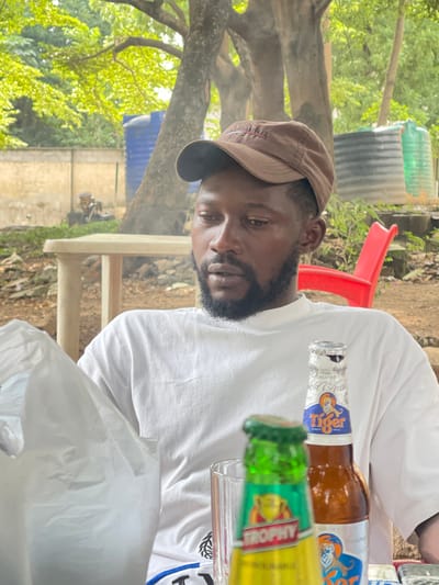 This image shows a man wearing a brown baseball cap and a white t-shirt, with a beard and a mustache. He is looking down and to the side. There are two beer bottles visible: one with a label that says "Tiger" and another with a label that says "Trophy." In the background, there are trees, some large blue and green water tanks, and a red plastic chair. The scene appears to be outdoors in a somewhat informal setting, possibly in Nigeria given the location context. The lighting suggests it might be daytime, with natural light filtering through the trees. The man's expression is difficult to discern fully due to the angle, but he does not appear to be smiling. The presence of beer bottles suggests a social gathering or relaxation. A white plastic bag is prominently in the foreground.