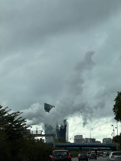 The image captures a view in Charenton-le-Pont, France, under a predominantly cloudy, overcast sky. Several modern, high-rise buildings are visible through a plume of what appears to be steam or smoke. A highway is present, with cars and traffic signs indicating the direction to Ivry-sur-Seine. Vegetation, including trees and bushes, frames the lower portion of the view.
